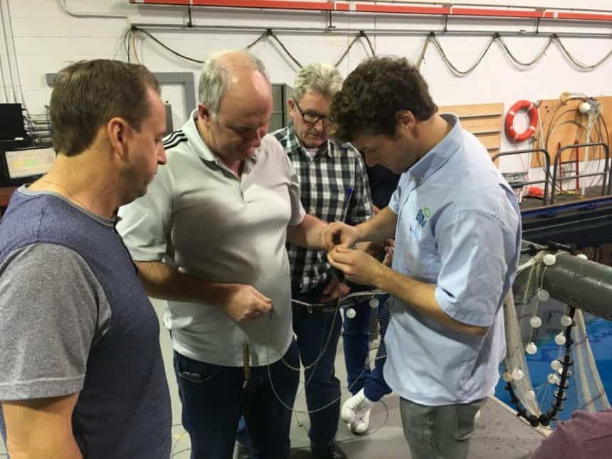Fishermen and industry experts examining fishing gear on a commercial vessel in a workshop setting.