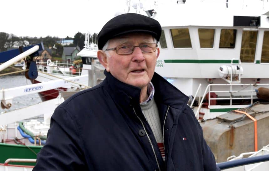 Experienced fisherman in front of fishing vessels at port, industry equipment visible in background.