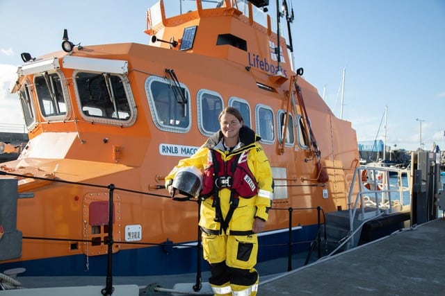 Commercial fishing vessel with RNLI crewwoman in safety gear at port, industry equipment visible.