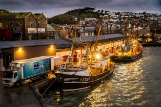 Fishing vessels docked at Newlyn port during evening hours, industry equipment visible in the scene.