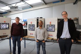 Three men in a marine engineering workshop with industrial equipment and control panels, commercialf.