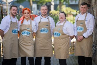 Young chefs at a seafood event, wearing aprons with 'Taste the Atlantic' logo, outdoors in a port or.