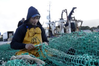 Fisherman inspecting fishing gear on a commercial fishing vessel in port, industry equipment visible.