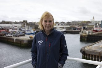 Female crew member on a fishing vessel at port, industry gear visible in background.
