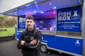 Commercial fishing vessel with Fish Box Dingle branding and crew member in front, industry equipment.