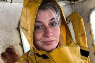 Fisherwoman in yellow rain gear on fishing vessel, industry equipment visible in background.