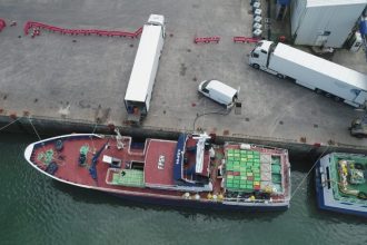 A fishing vessel docked at port with industry equipment and cargo containers visible.