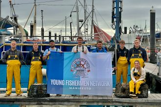 Young fishermen with a banner at a busy fishing port, industry equipment visible in background.