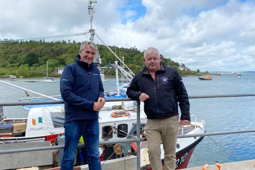 Two fishermen stand on dock with fishing vessel, industry gear, port background, industry focus, noY.