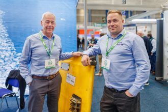 Two fishermen at a trade show with industry equipment in a commercial fishing industry setting.