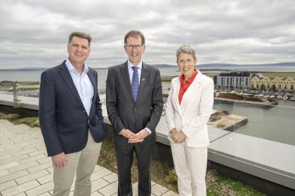 Three professionals standing outdoors with Galway Bay in background during cloudy day.