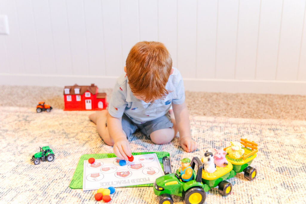 A young boy with ginger hair is kneeling on the floor. He is playing with a farm prepositions game and there are farm toys scattered around him.