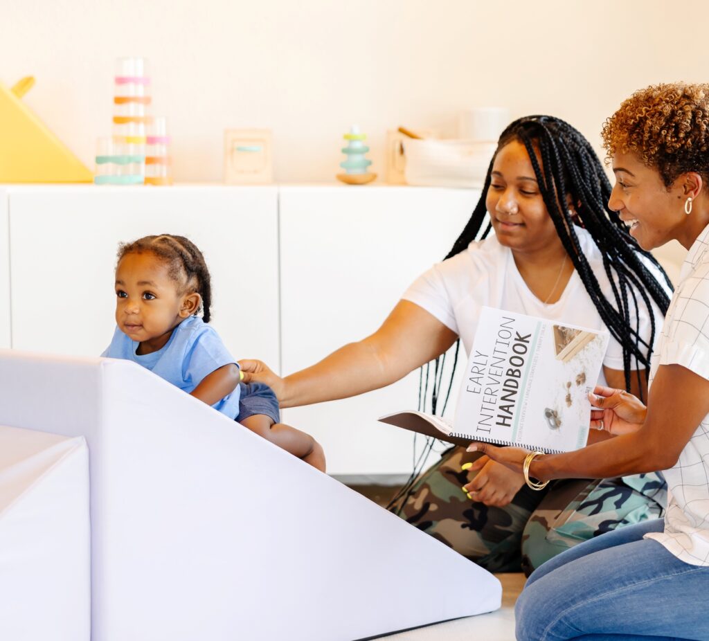 Caregiver Coaching example - picture of a therapist speaking to a mother while her son plays on a foam block. The therapist is holding a copy of The Early Intervention Handbook.
