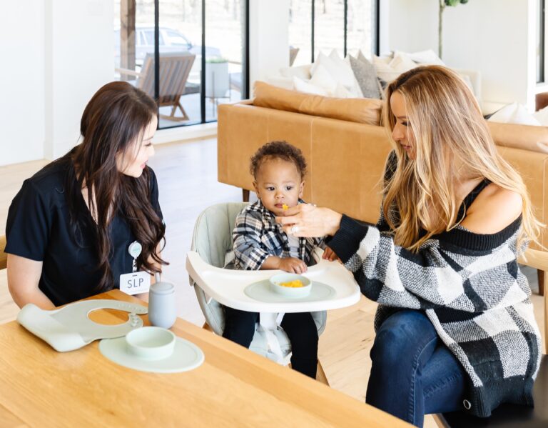 Image of a therapist sat at a dining table. There is a baby in a high chair and the mum is next to the baby, feeding him a snack.