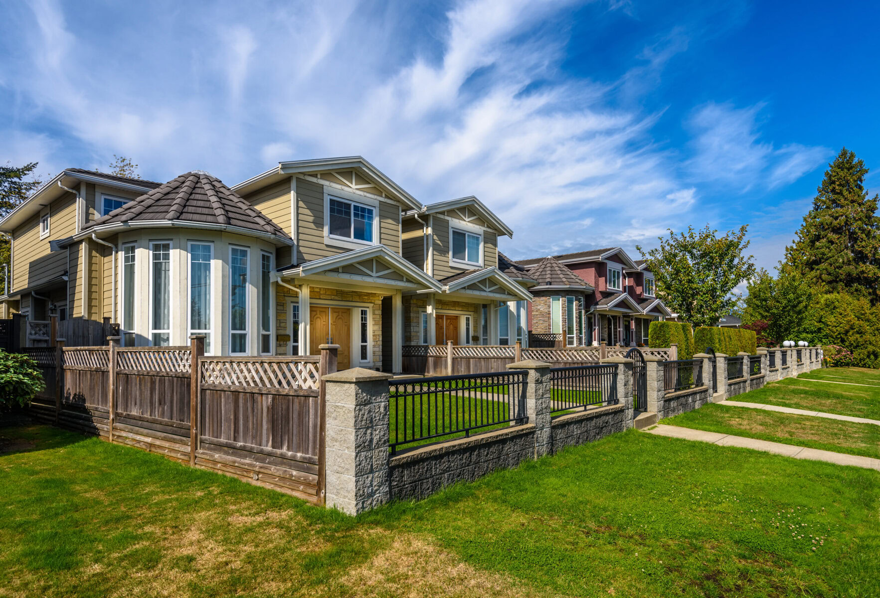 Row of houses with fences and green lawns under a blue sky.