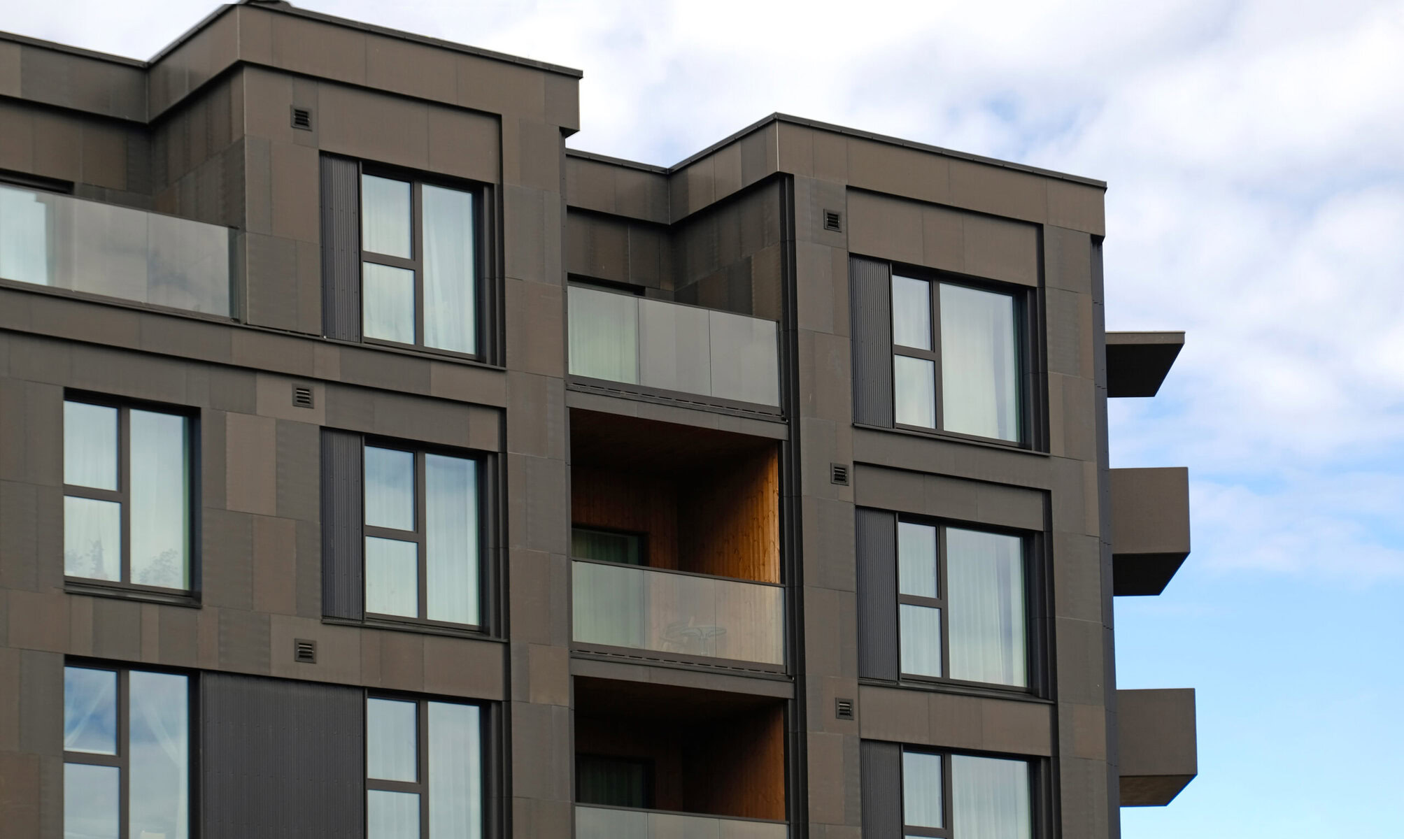 Modern apartment building with dark facade and glass balconies against a cloudy sky.