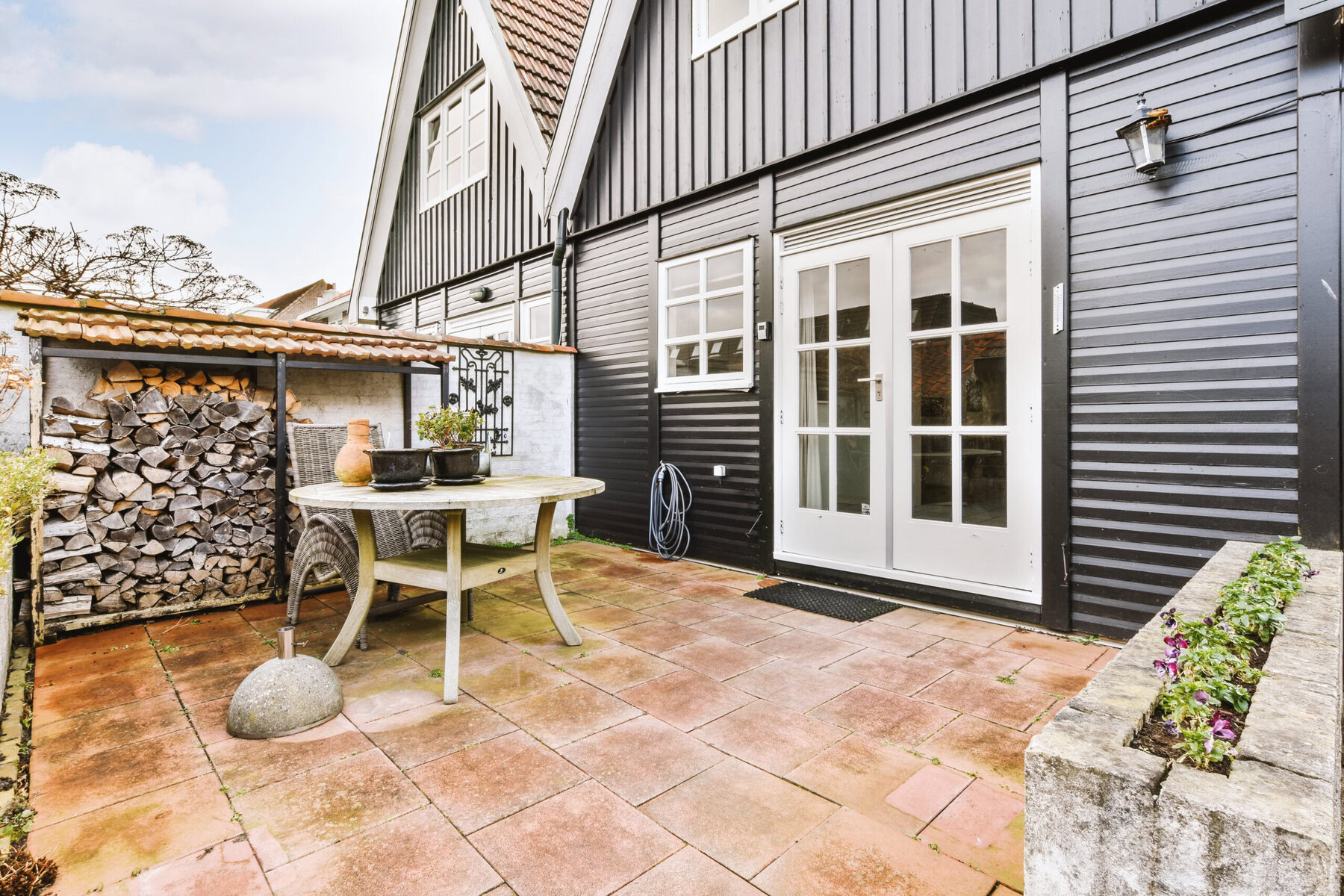 Outdoor patio with a white table, chairs, and stacked firewood next to a dark house.