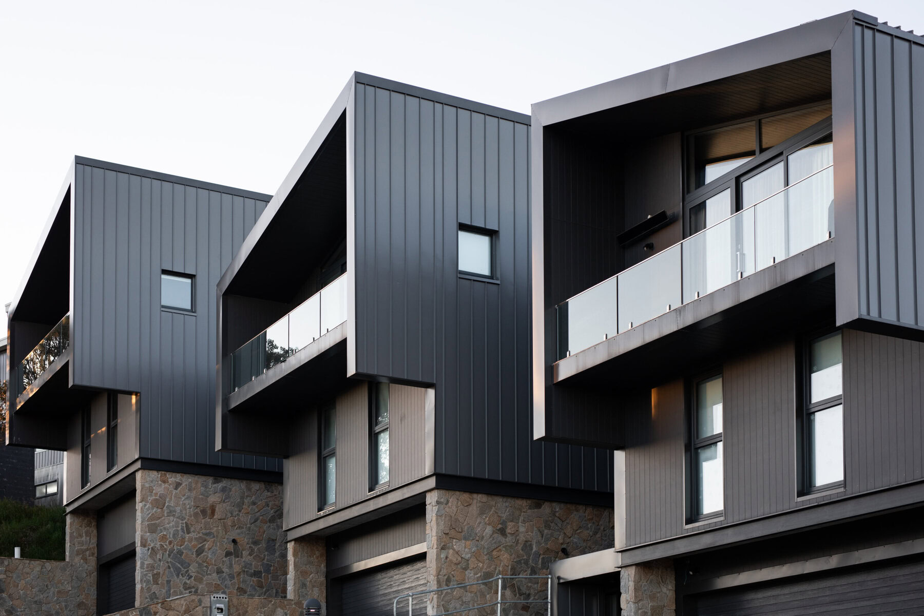 Modern dark gray townhouses with stone bases and glass balconies under a clear sky.