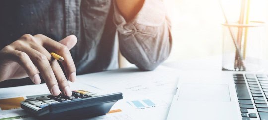 Person calculating finances with calculator and laptop at desk.