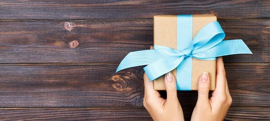 Person holding a gift box wrapped with a blue ribbon on wooden background.
