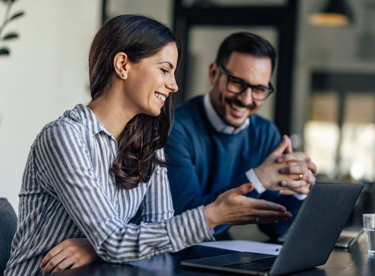 2 people smiling and looking at a laptop while sitting next to each other
