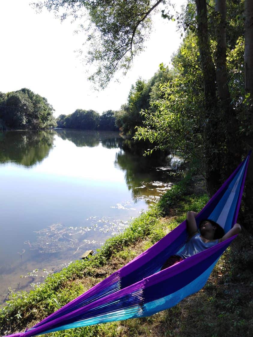 A girl relaxing in a purple blue hammock tied between trees next to a clear river.