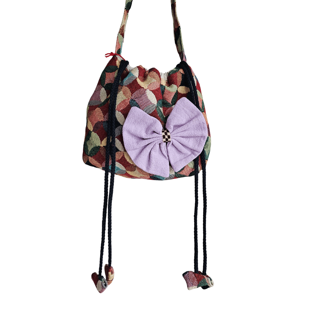 A stylish drawstring bucket bag or pouch bag featuring a vibrant, multi-color geometric fabric pattern with interlocking circle or Seigaiha wave motifs with bohemian style. The textile includes shades of teal, burgundy, olive green, cream, and brown. Decorated with purple bows and wood beats