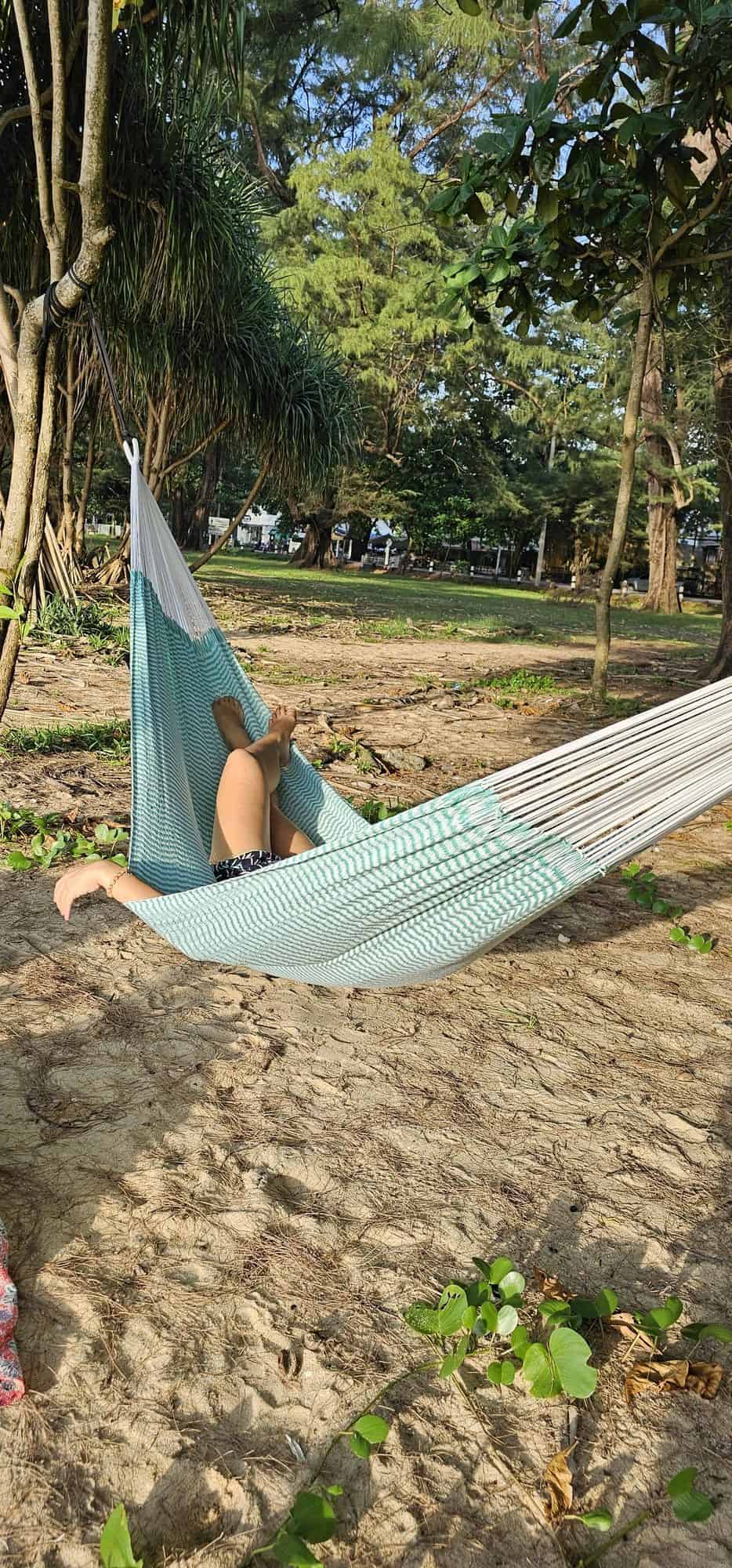ULA "Mixcloud Mint" hammock in a mint green and white chevron pattern, with white ropes, relaxing on a sandy, treed beach or tropical park area.