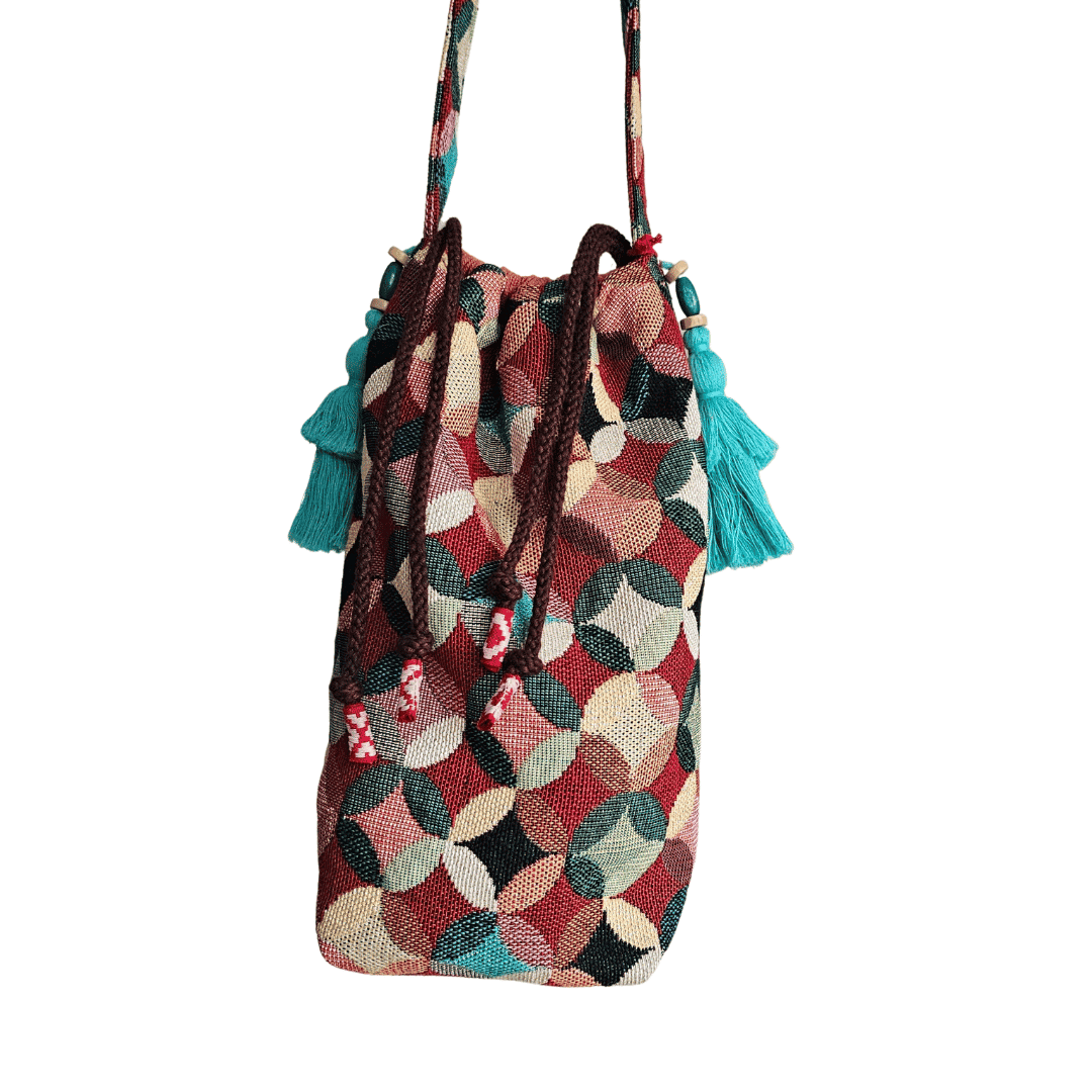 A stylish drawstring bucket bag or pouch bag featuring a vibrant, multi-color geometric fabric pattern with interlocking circle or Seigaiha wave motifs with bohemian style. The textile includes shades of teal, burgundy, olive green, cream, and brown. Decorated with tassels and wood beats
