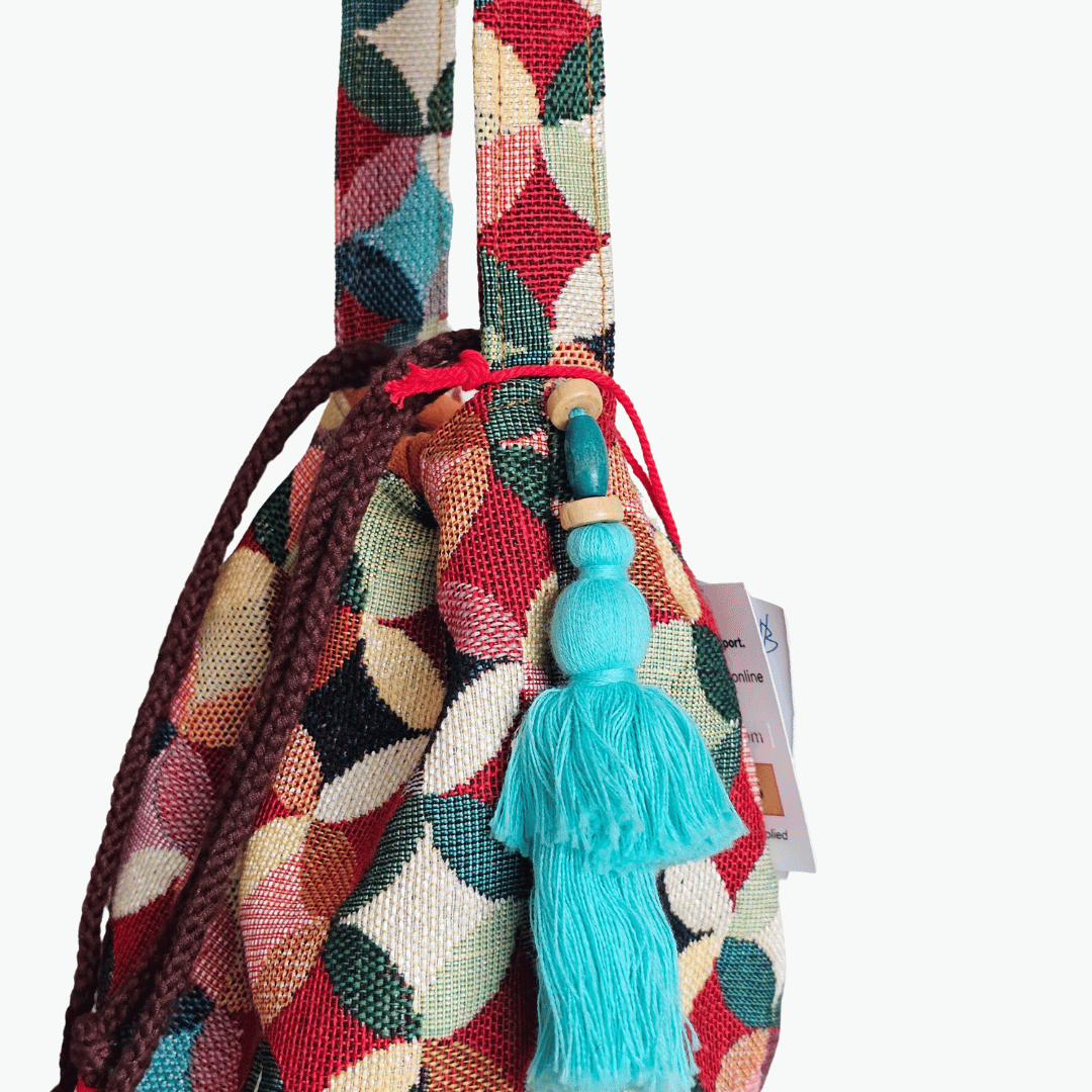 A stylish drawstring bucket bag or pouch bag featuring a vibrant, multi-color geometric fabric pattern with interlocking circle or Seigaiha wave motifs with bohemian style. The textile includes shades of teal, burgundy, olive green, cream, and brown. Decorated with tassels and wood beats