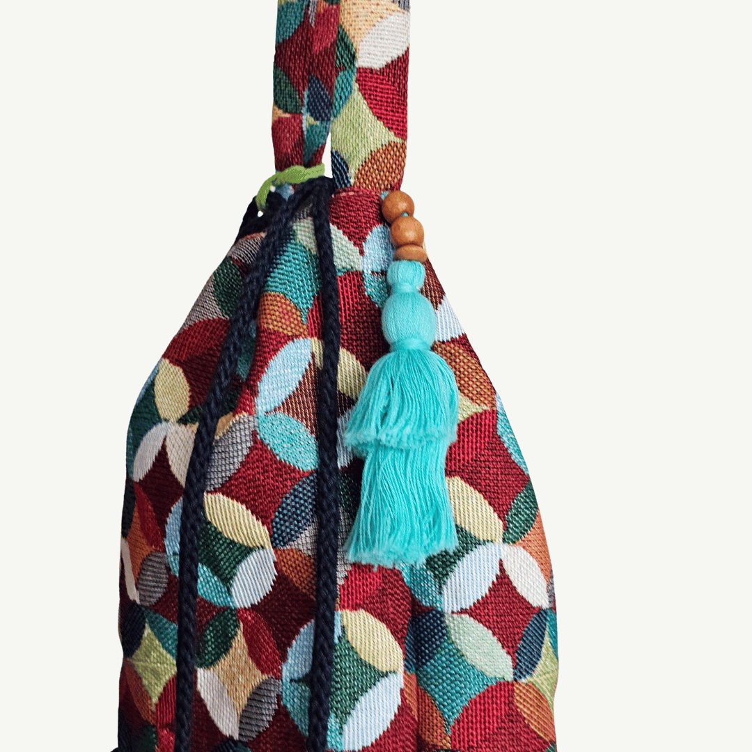 A stylish drawstring bucket bag or pouch bag featuring a vibrant, multi-color geometric fabric pattern with interlocking circle or Seigaiha wave motifs with bohemian style. The textile includes shades of teal, burgundy, olive green, cream, and brown. Decorated with tassels and wood beats