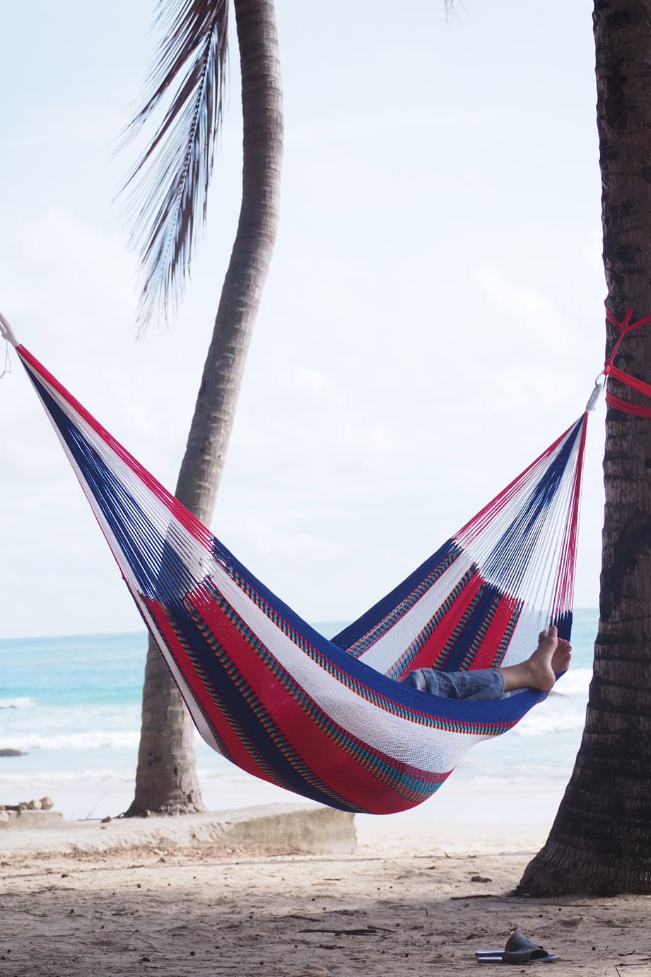 Person relaxing in a multi-colored Thailand-pattern acrylic yarn hammock, hung between two palm trees on a tropical beach near the ocean.