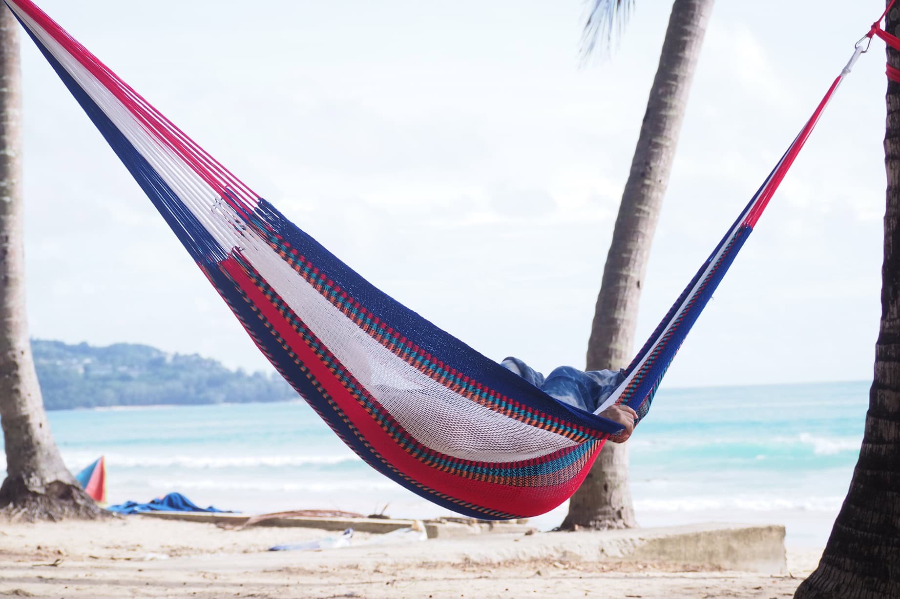 Person relaxing in a multi-colored Thailand-pattern acrylic yarn hammock, hung between two palm trees on a tropical beach near the ocean.