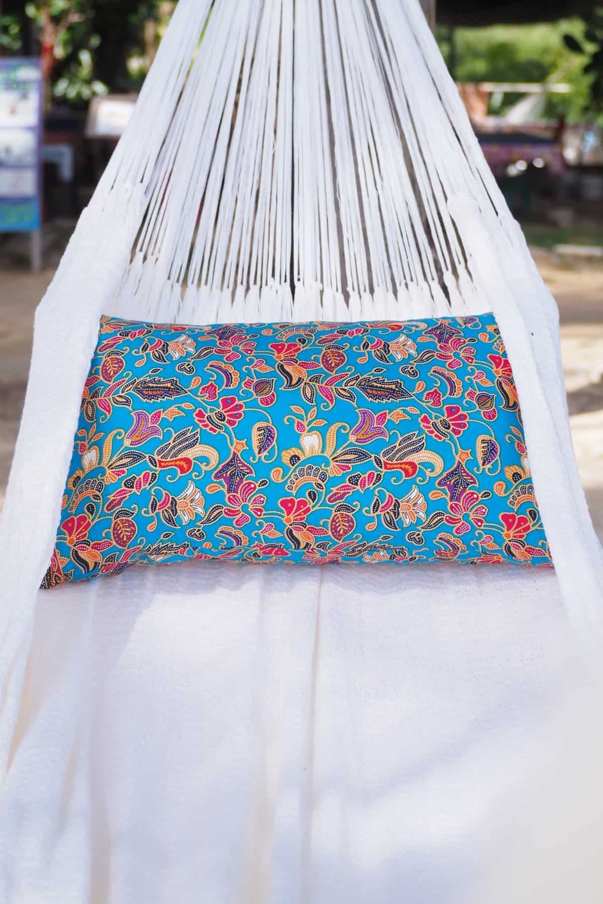 Close-up of a pure white ULA hammock hanging from a palm tree, accessorized with a vibrant blue, red, and yellow floral pillow.