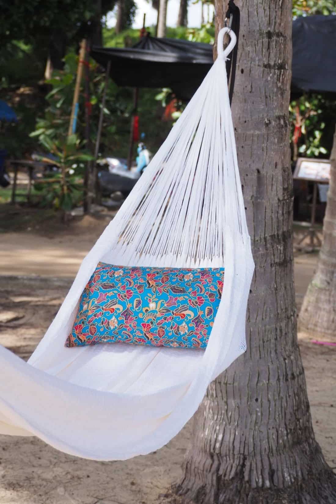 Close-up of a pure white ULA a pure white ULA hammock hanging from a palm tree, accessorized with a vibrant blue, red, and yellow floral pillow.Travel hammock hanging from a palm tree, accessorized with a vibrant blue, red, and yellow floral pillow.