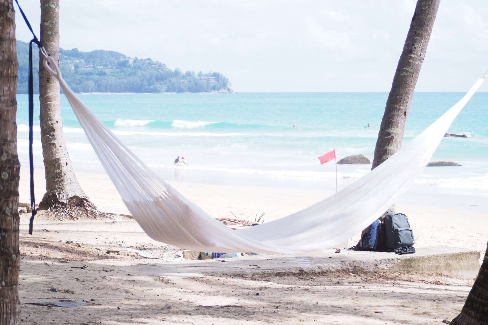a pure white ULA hammock hanging from a palm tree, accessorized with a vibrant blue, red, and yellow floral pillow.