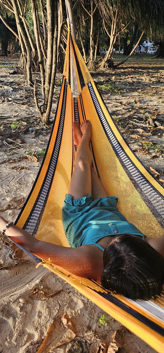 Close-up of a brightly striped yellow and navy blue rope hammock, tied between trees on a sandy coastline at sunset.