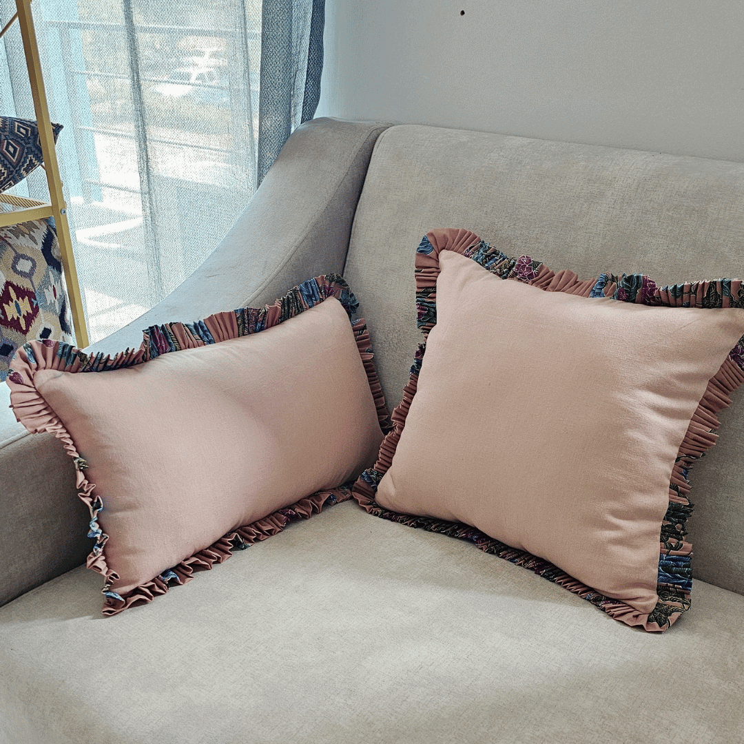 Two rectangular lumbar throw pillows with a dusky pink blush cotton body and a wide, ruffled trim featuring a batik cotton floral fabric edge. The batik fabric displays a vibrant, multicolor pattern in shades of teal, purple, fuchsia, and navy. The decorative cushions are resting on a neutral gray sofa, blending soft cotton texture and eclectic color for stylish home decor and interior design emphasis.