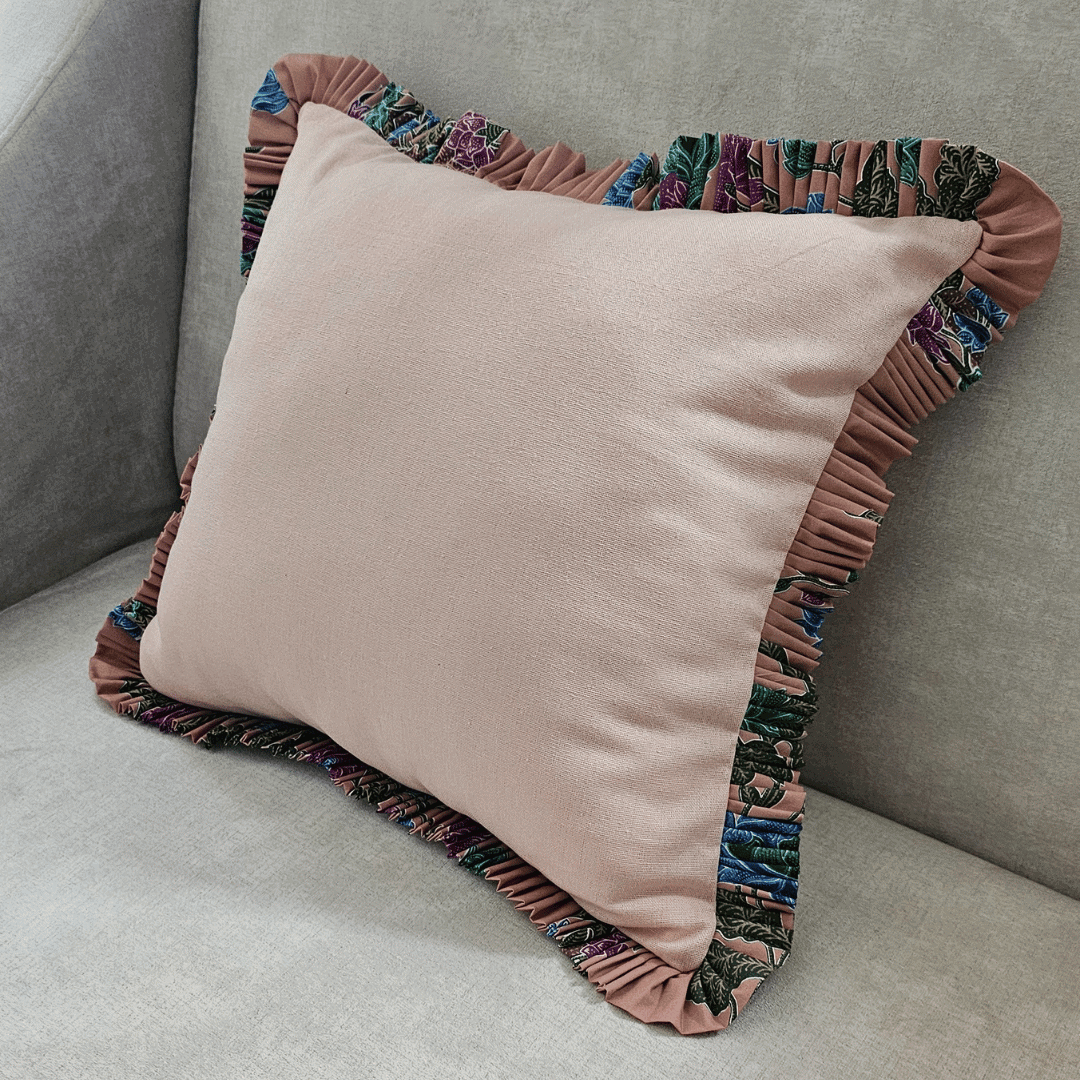 A square throw pillow featuring a dusky pink blush cotton body with a wide, decorative ruffled trim. The ruffle is detailed with a vibrant multicolor batik cotton floral fabric in shades of teal, purple, fuchsia, and navy. This decorative cushion is resting on a neutral gray sofa, highlighting a blend of soft texture and eclectic home decor style for a living room.