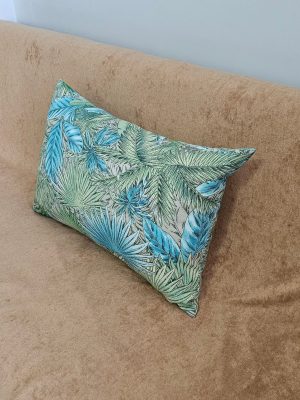 A throw pillow featuring a vibrant tropical leaf print or jungle botanical pattern in shades of teal, turquoise, and various greens. The decorative cushion is placed on a tan or camel-colored sofa with a suede-like texture, set against a light gray wall. This home decor accent provides a bright pop of color and a maximalist or eclectic tropical style for a living space.