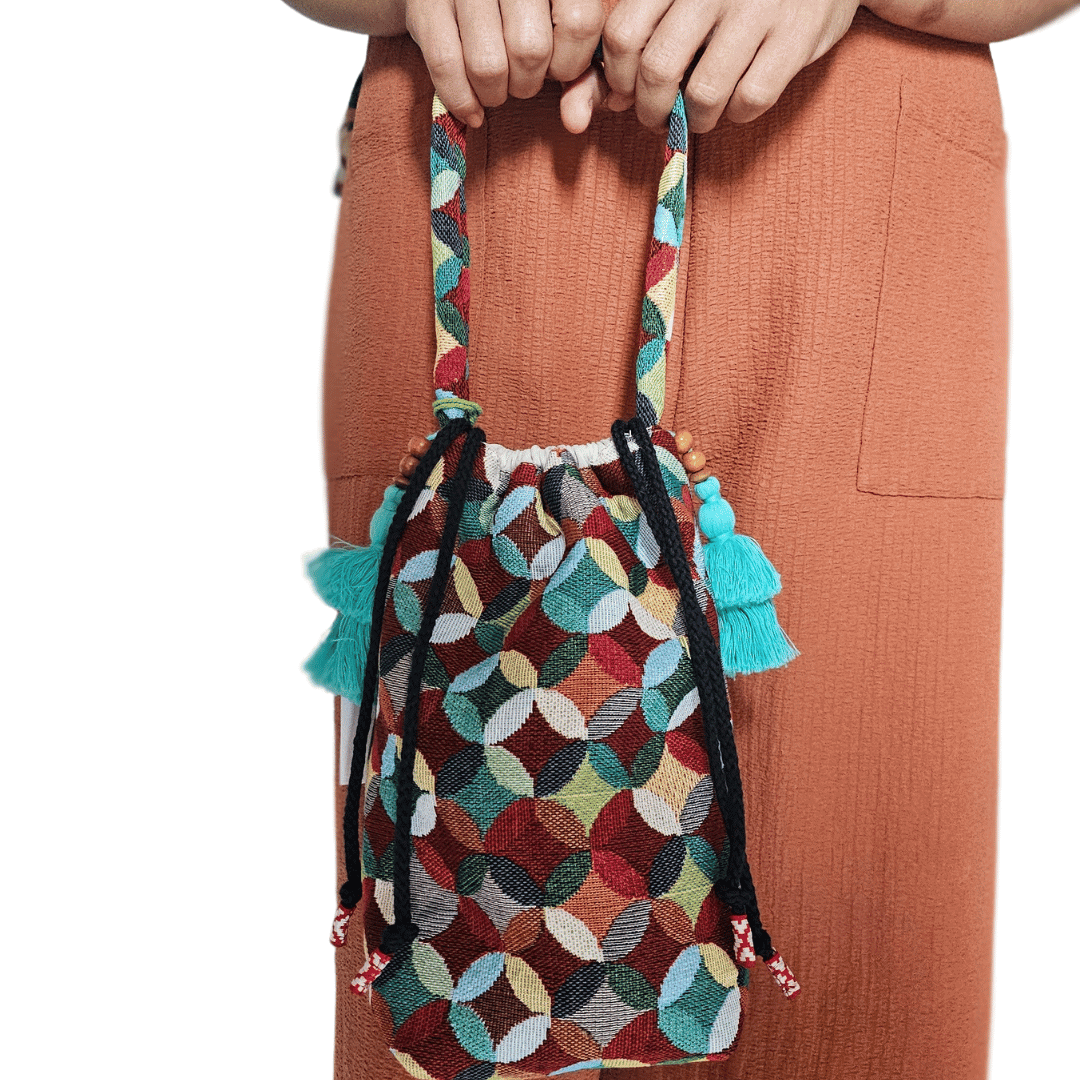 A stylish drawstring bucket bag or pouch bag featuring a vibrant, multi-color geometric fabric pattern with interlocking circle or Seigaiha wave motifs with bohemian style. The textile includes shades of teal, burgundy, olive green, cream, and brown. Decorated with tassels and wood beats