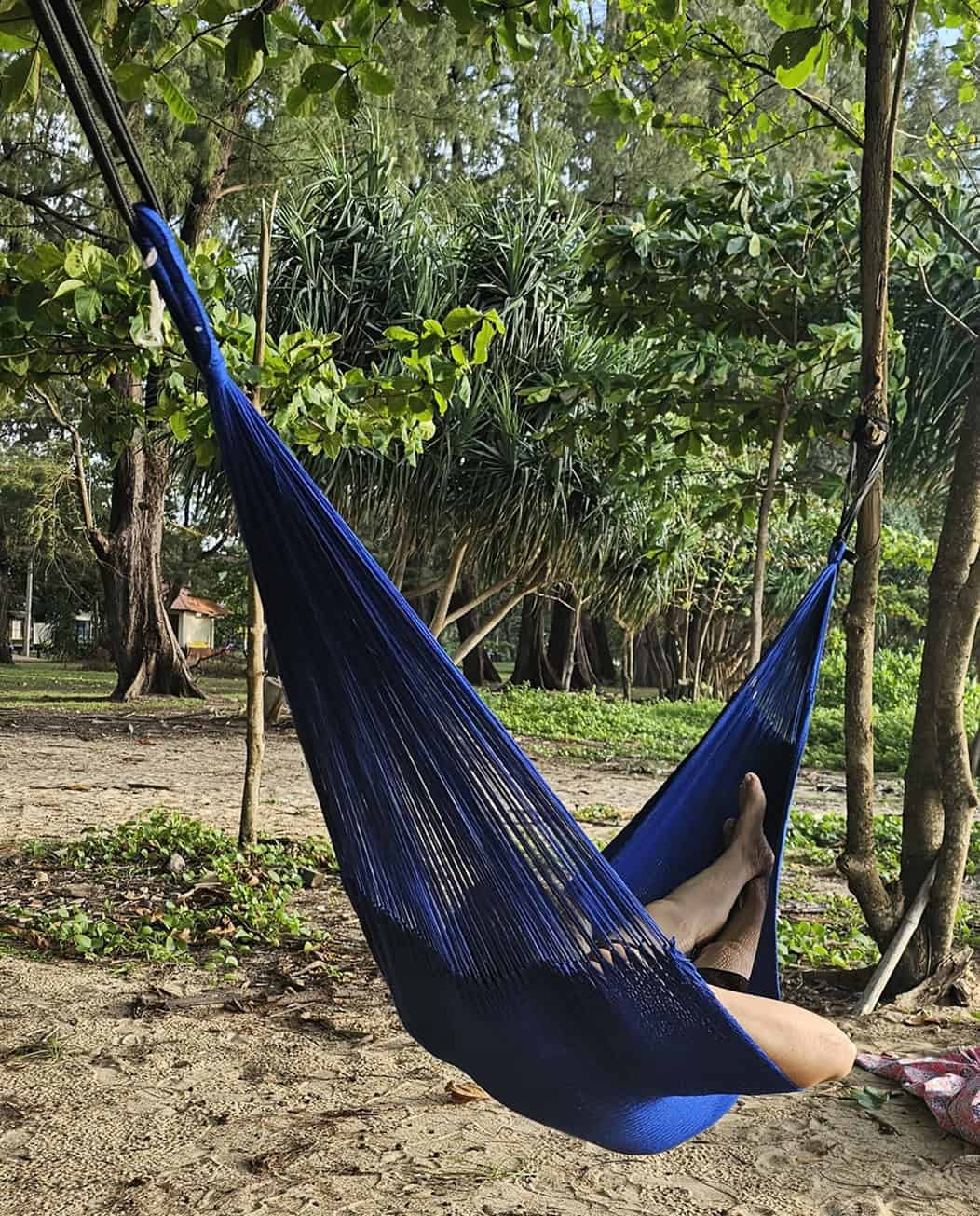 "Royal Blue" hand-woven hammock, gathered-end style, featuring a person relaxing on a sandy beach under palm trees.