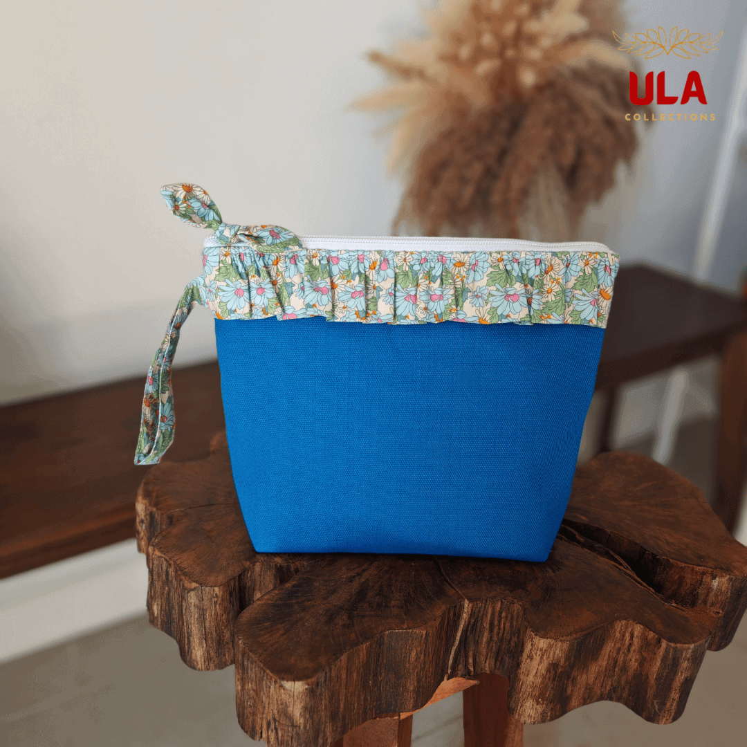 Front shot of a blue canvas ULA Collections pouch with a light blue and peach floral ruffled trim and a coordinated bow zipper pull.
