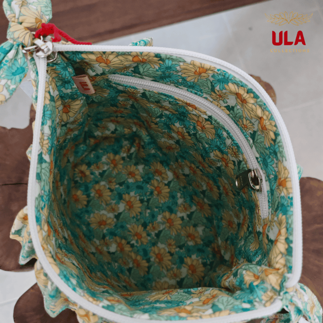 Close-up angled view of an open blue pouch, highlighting the white zipper, green floral ruffles, and the ULA brand tag inside.