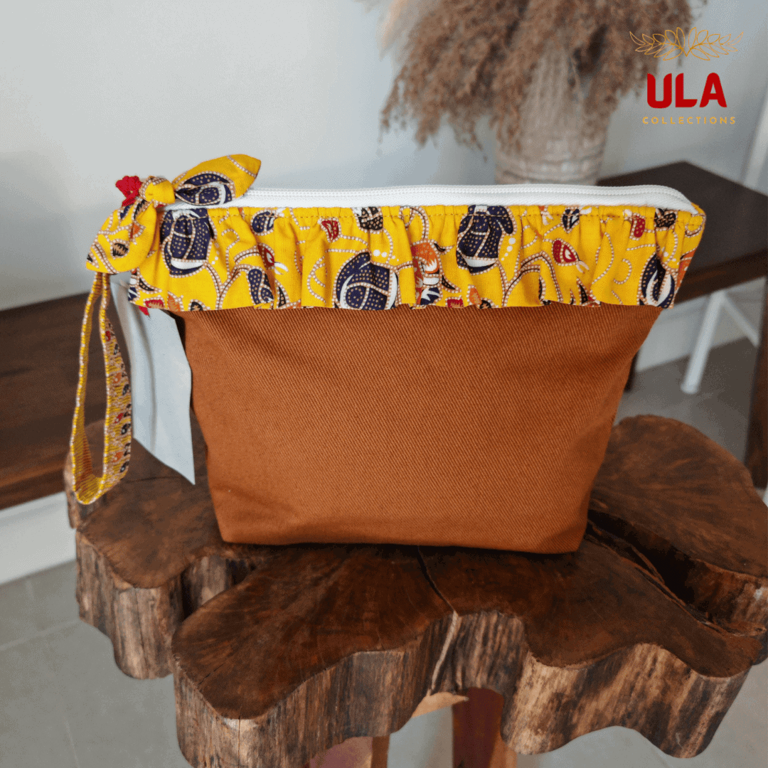 A handcrafted brown canvas pouch by ULA Collections, featuring a vibrant yellow Batik print ruffled trim and matching wristlet strap, displayed on a natural wood stand.