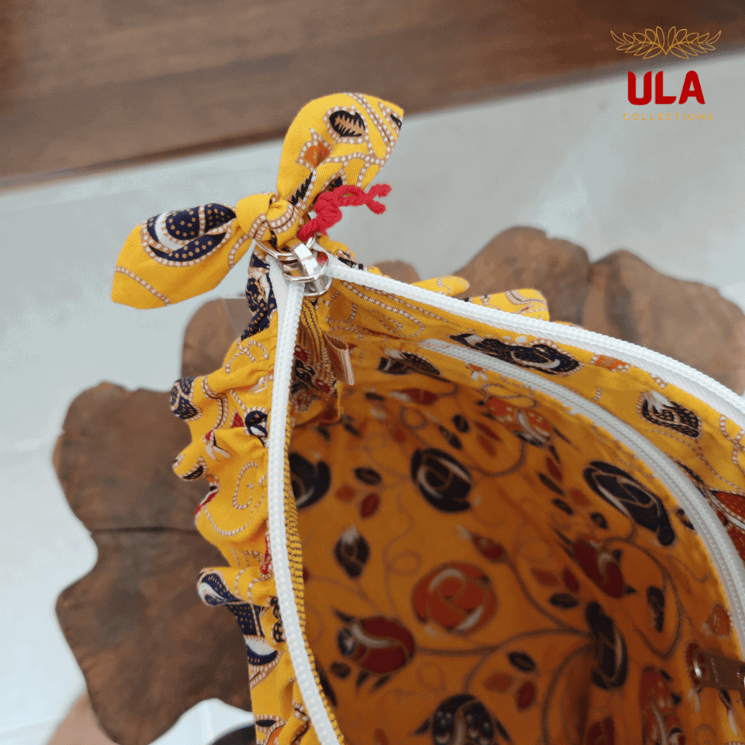 Close-up of a white zipper on a yellow cotton patterned toiletry bag, showing a decorative fabric bow and the internal ULA brand tag.