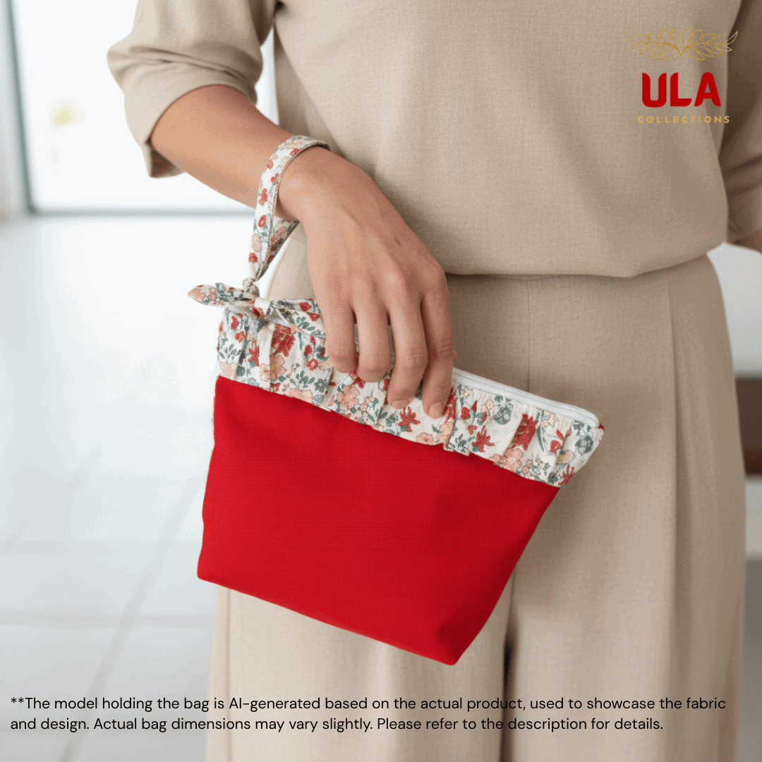 An AI-generated lifestyle image of a model wearing a beige outfit, holding a red canvas pouch. This bag is accented with a cream-colored floral ruffle featuring delicate red and green botanical patterns. The model holds the bag by its matching patterned wristlet strap. The background is a bright, minimalist indoor space, and the "ULA COLLECTIONS" logo appears in the corner.