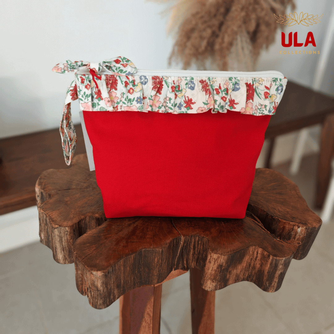 Front view of a vibrant red canvas cosmetic pouch featuring a ruffled floral trim and a matching bow zipper pull, displayed on a rustic wooden stool.