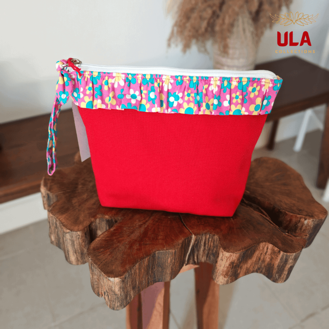 Front view of a vibrant red canvas pouch from ULA Collections, featuring a pink ruffled top trim with a teal and yellow floral pattern, placed on a decorative wooden stand.