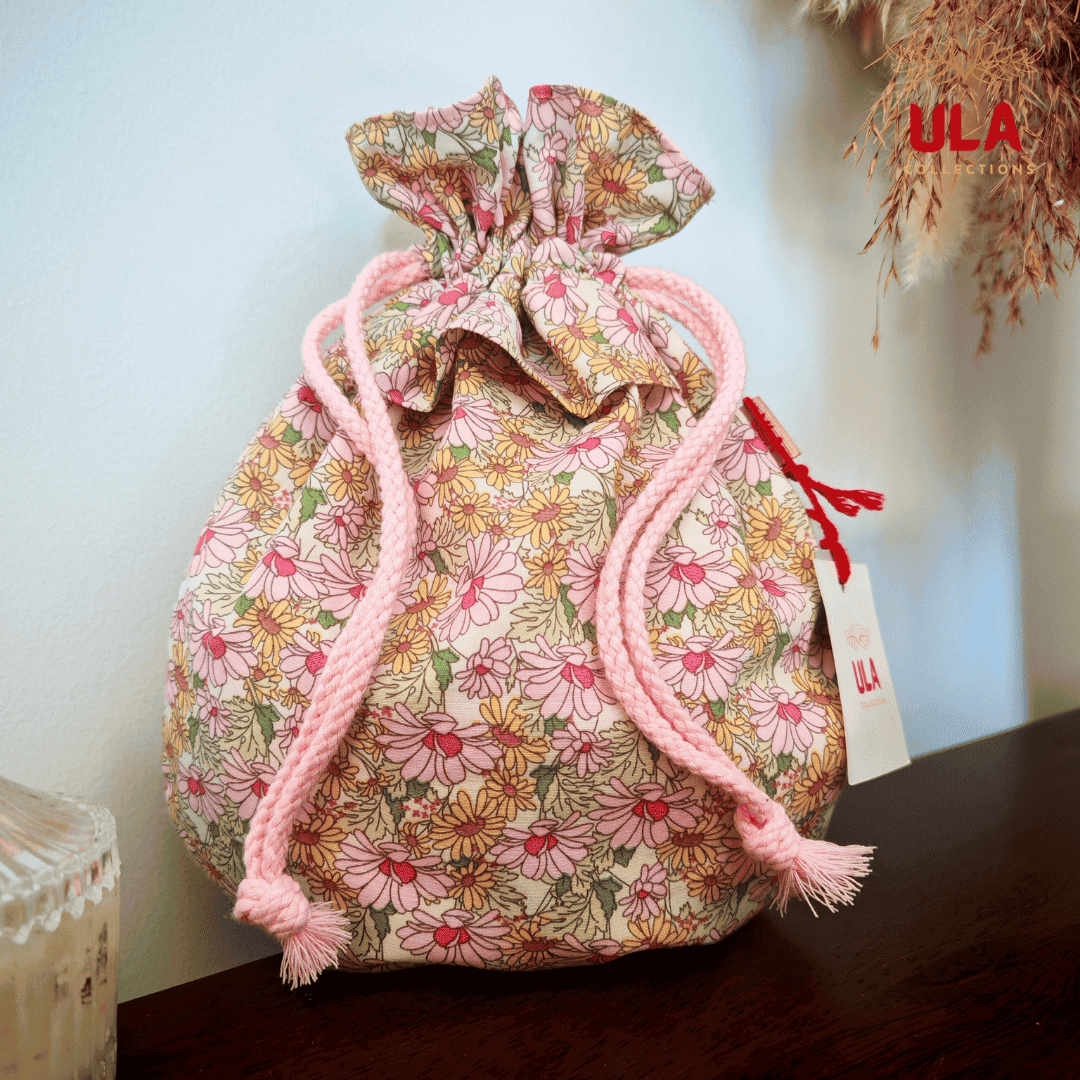 A full front view of a drawstring bag featuring a dense daisy floral pattern in shades of pink, yellow, and green. The bag has soft pink braided cords and an accent flap of blue and pink batik-style fabric tucked at the top.